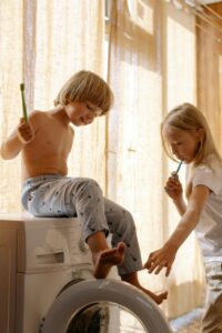 Two kids brushing teeth indoors on washing machine in natural light.