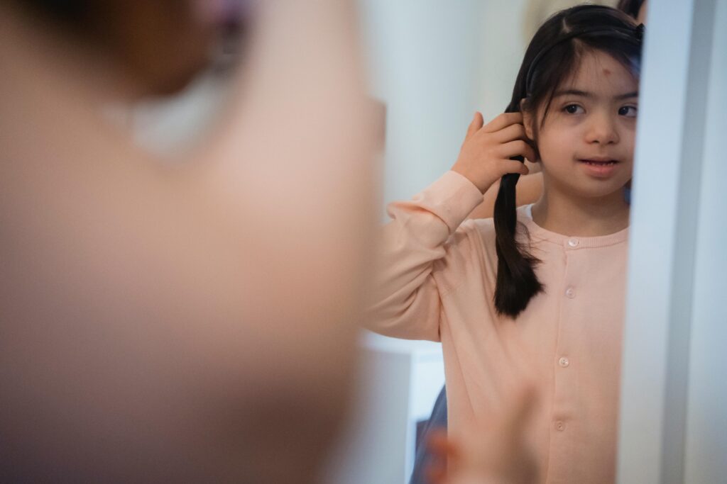 A young girl with long hair adjusts her hairstyle in front of a mirror indoors.