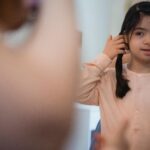 A young girl with long hair adjusts her hairstyle in front of a mirror indoors.